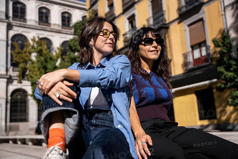 A lesbian couple enjoys a sunny day in a European city, sitting casually and soaking in the warm atmosphere. Dressed stylishly with sunglasses, they embrace the moment with confidence and joy
