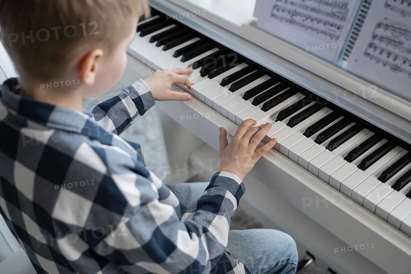 A young child wearing a plaid shirt is focused on playing the piano, their fingers poised over the keys, with open music sheets placed in front of them on the piano stand