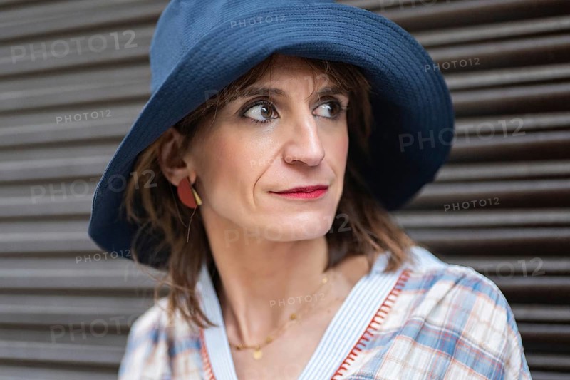 A fashion woman wearing a blue hat and plaid shirt stands against a dark background, exuding a thoughtful and stylish aura. Her red earrings and makeup add a pop of color