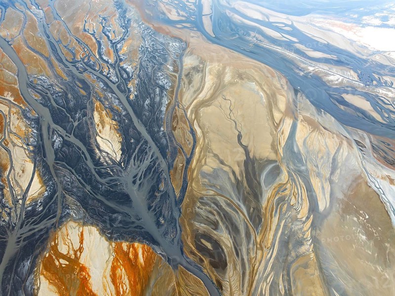From above, this mesmerizing aerial capture reveals intricate patterns created by river systems, resembling an abstract painting with a blend of vivid earth tones and cool blues that weave through the landscape