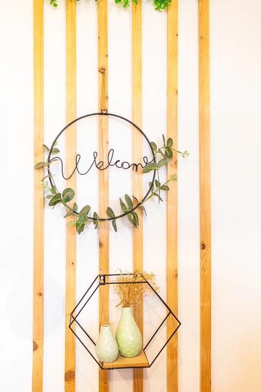 An elegant decorative setup featuring a metal Welcome sign adorned with greenery Below, modern ceramic vases are displayed on a geometric shelf against vertical wooden slats