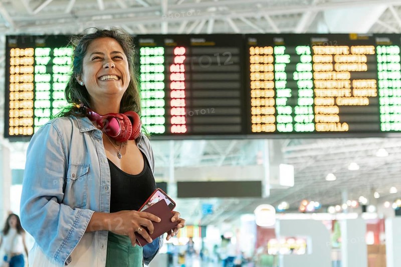 A woman at an airport smiles joyfully, headphones around her neck and holding a passport The flight information board in the background enhances the travel excitement
