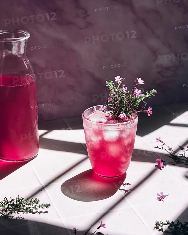 A glass of vibrant pink lemonade, garnished with delicate purple flowers, sits beside a transparent pitcher. Sunlight filters in, casting artistic shadows on the tiled surface