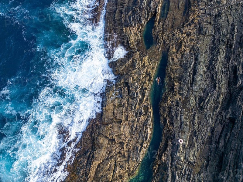 Top view of natural rock pools in Tindaya, Fuerteventura, featuring a woman leisurely swimming in clear blue waters, surrounded by rugged cliffs and waves crashing nearby
