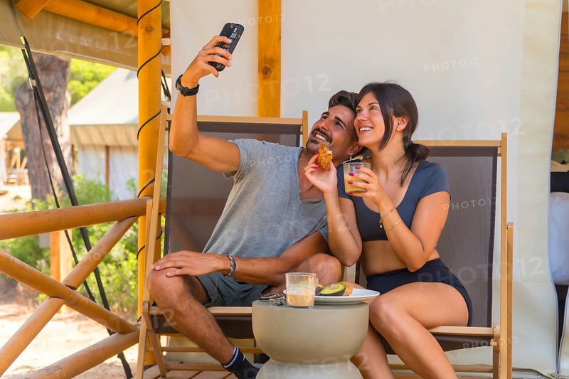Happy couple capturing a selfie with their smartphone while savoring a healthy breakfast on the wooden veranda of their luxurious glamping tent, enjoying a peaceful summer morning together