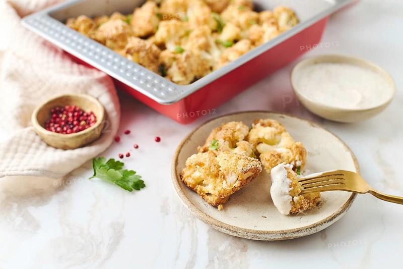Baked cauliflower coated with breadcrumbs and spices in a red metal form. The cauliflower is served with sauce in a cozy kitchen setting, alongside ingredients like parsley and spices, highlighting a homemade, healthy meal