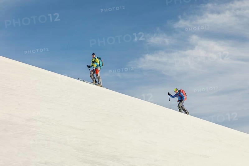 Two skiers with backpacks ascend a snowy mountain slope under a clear blue sky, showcasing the adventure and sport of ski touring in a winter wonderland