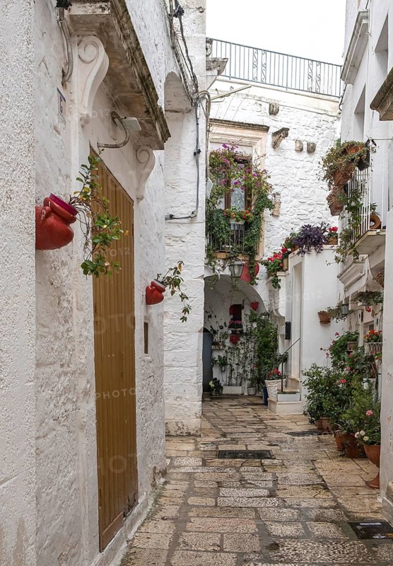 Narrow Mediterranean alleyway with white buildings, decorated with flowering plants and rustic doors, Cisternino, Apulia, Italy