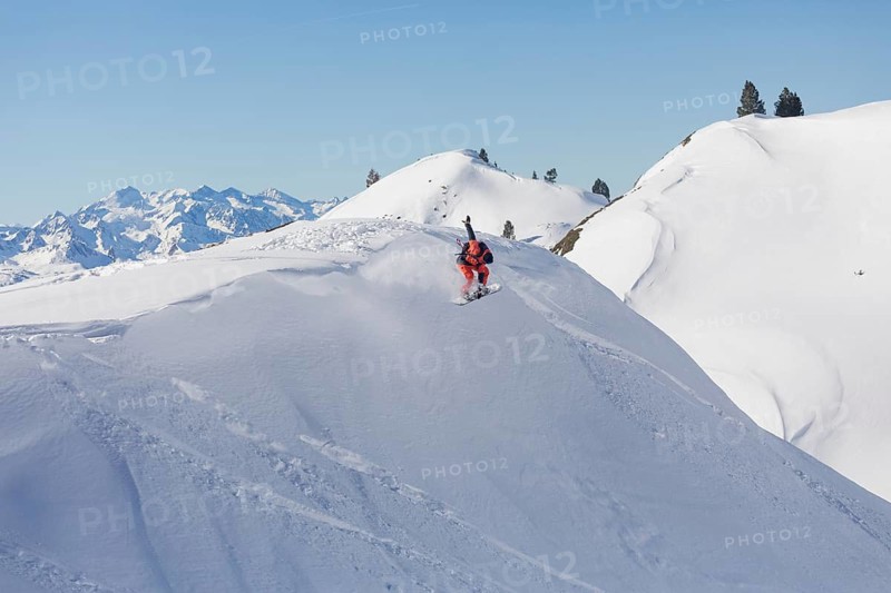 A snowboarder in a red jacket executes a dynamic aerial trick on a snow covered mountain slope, surrounded by scenic, snowy peaks under a clear blue sky