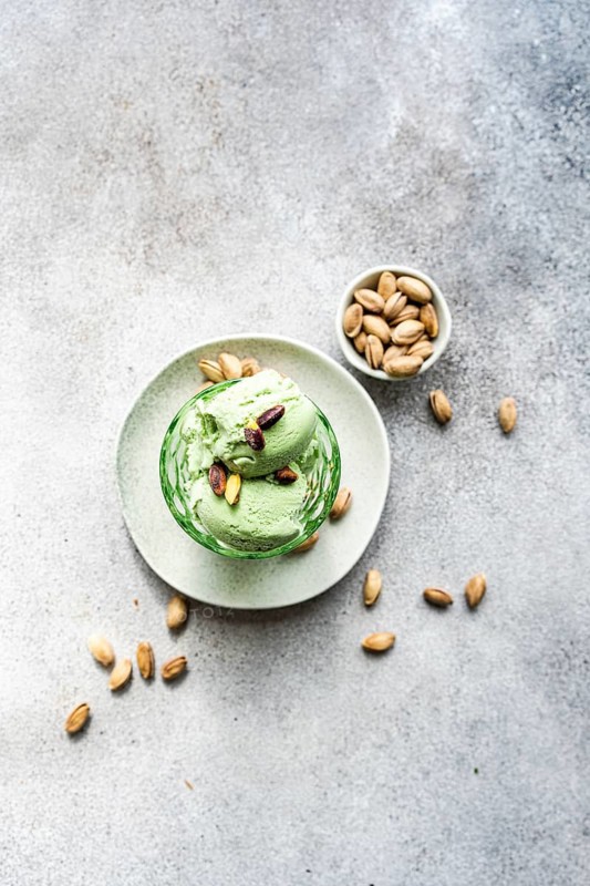 A creamy and homemade scoop of pistachio ice cream sits in a green bowl, surrounded by whole pistachios scattered artfully on a textured gray surface. Perfect for dessert themed visuals