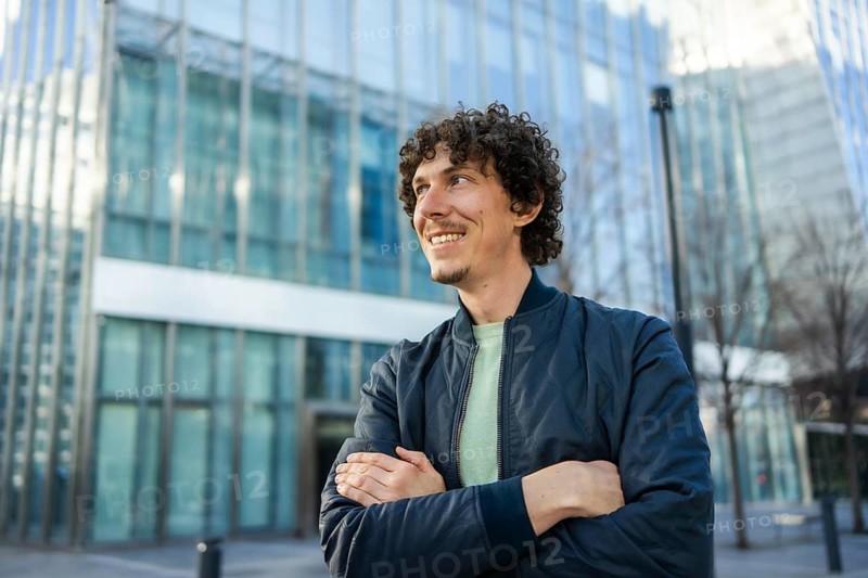 A cheerful man stands confidently with crossed arms, smiling in an urban setting. The background features a modern glass building, conveying a sense of business and city life