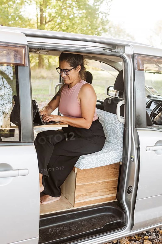 A woman works remotely from a cozy van in the scenic autumn setting of New Zealand. She is focused on her laptop, surrounded by colorful foliage and nature