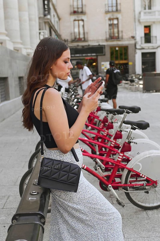 A young woman stands by a row of rental bicycles, texting on her phone. She is dressed casually for a day out in the city, with a purse slung over her shoulder