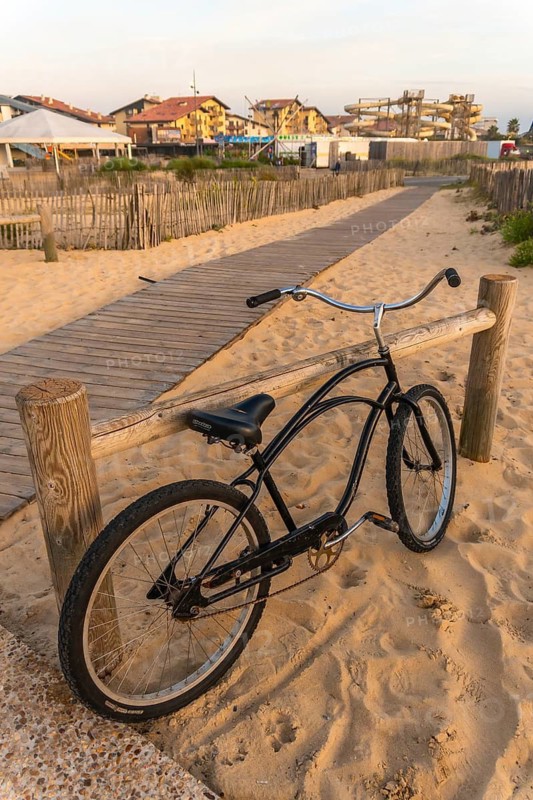 Bicycle leaning against a wooden fence on a sandy path leading to beach town buildings and water park structures, capturing a sunny summer holiday scene