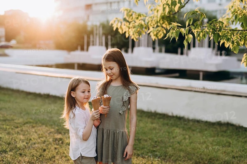 Two young girls enjoy ice cream cones on a sunny day in a park of the city. Surrounded by greenery and soft sunlight, they share smiles and joy in a peaceful outdoor setting