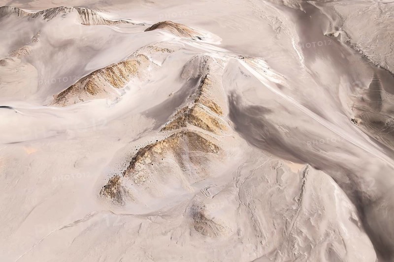 An aerial view captures the rugged desert landscape, showcasing rolling sand dunes and rocky formations. The natural patterns in the sand create a visually striking scene