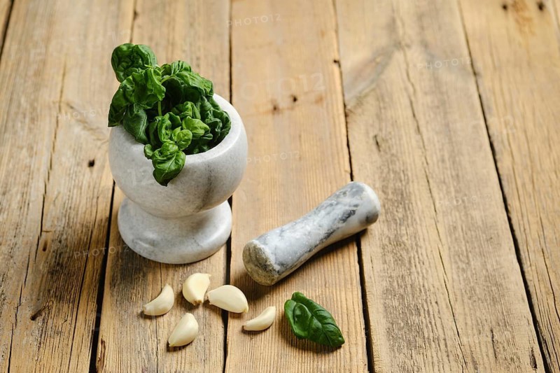 Basil leaves are placed in a mortar with a pestle beside it. Garlic cloves sit on the wooden table. This scene shows the preparation for making a recipe with fresh ingredients