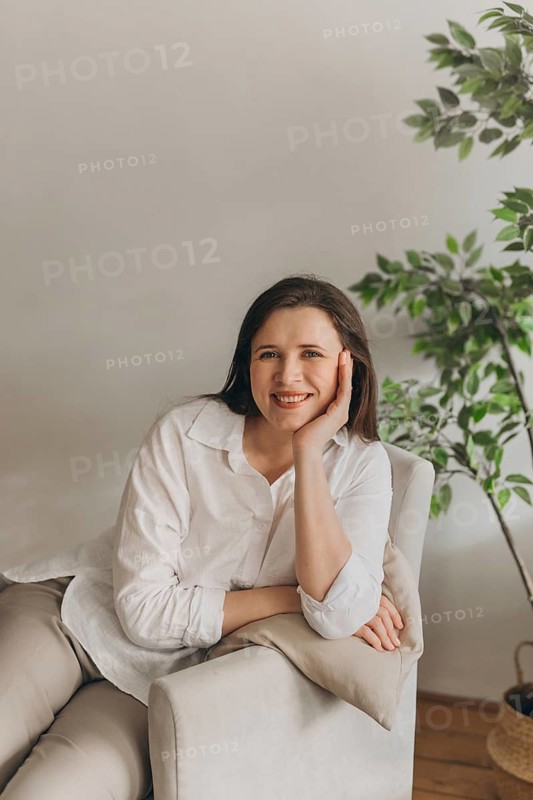 Smiling woman sitting casually on a chair in a cozy room with green plants. The relaxed atmosphere suggests a focus on wellness and nutrition. Ideal for lifestyle and health themes