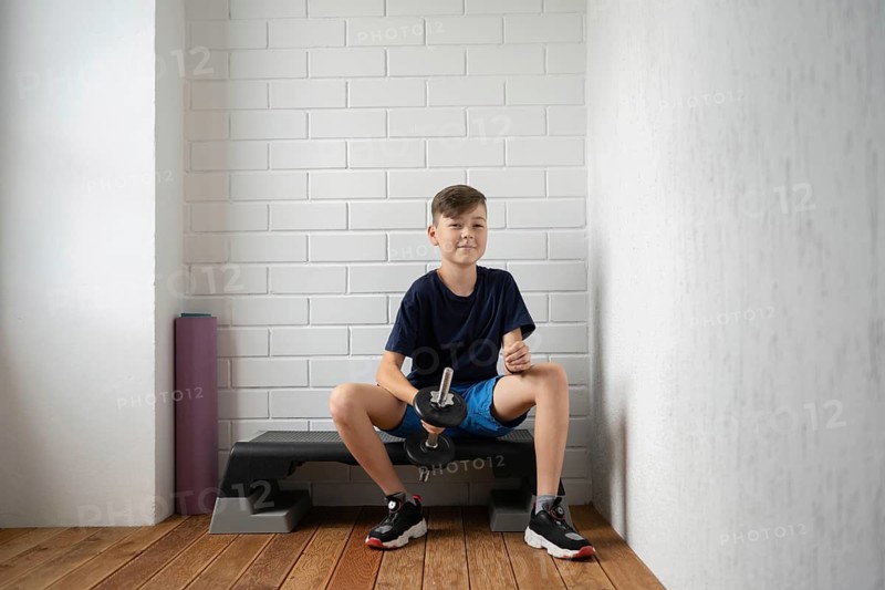 Young boy in blue shorts and t-shirt sits confidently on an exercise bench, holding a dumbbell in a home gym setting with a white brick wall background and a wooden floor