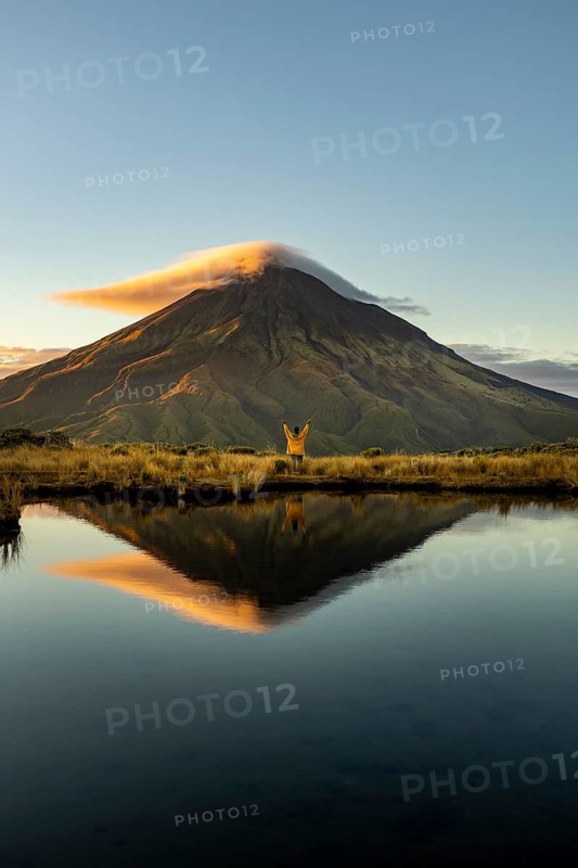 Back view of unrecognizable person with arms raised admiring the stunning sunrise reflection of Mount Taranaki in New Zealand. A serene moment captures the majestic nature and tranquility of this landscape