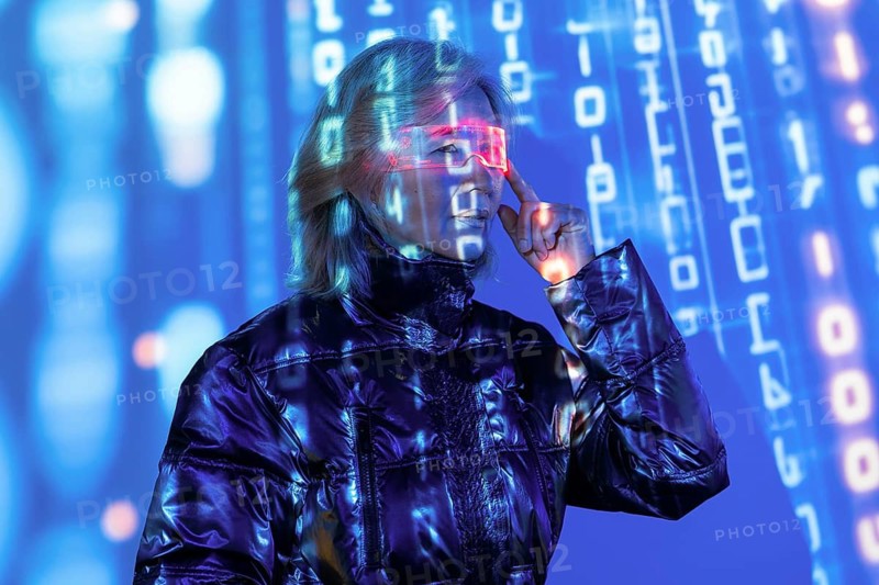 An elderly Asian woman engages with the metaverse, wearing futuristic glasses. She is surrounded by digital data and immersed in a virtual environment, showcasing modern technology