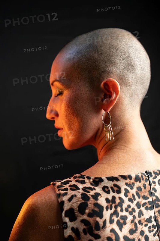 A bald woman is captured from the side wearing a leopard print dress and a gold earring. The lighting highlights her unique style and elegance against a dark background