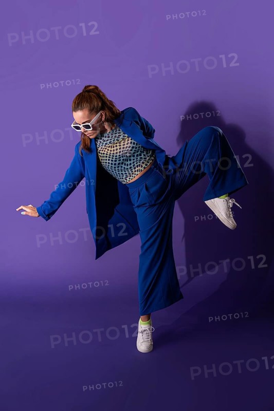 Young woman dancer striking a dynamic pose in a stylish blue outfit with white sneakers, set against a vivid purple background, showcasing modern fashion and expressive movement