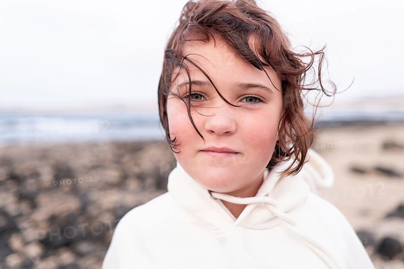 A young child with wind-swept hair gazes at the camera on a summer day by the beach. The ocean creates a tranquil background for this candid portrait of carefree youth