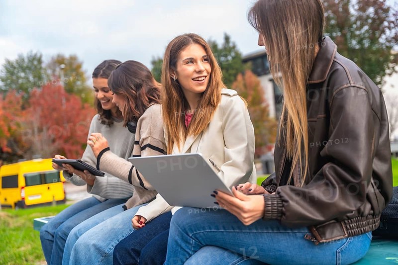 Group of young women friends studying together on campus, utilizing laptop and tablet for shared learning while sitting on a bench with autumn trees in the background