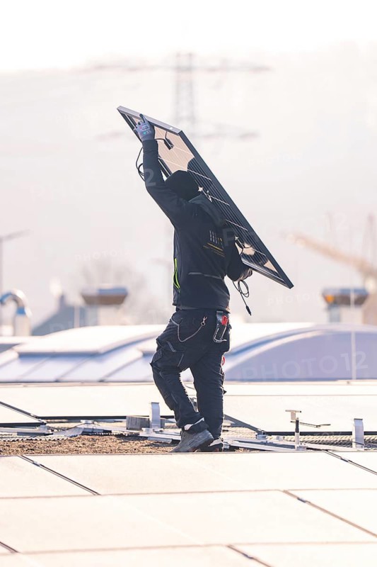 Worker in protective clothing lifts solar module on a sunny roof surrounded by urban landscape, PV installation at OBI Baumarkt, Mühlacker, Enzkreis, Germany