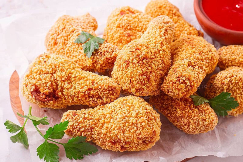 A wooden board holds several golden chicken drumsticks arranged neatly. A small bowl of red dipping sauce is nearby. Fresh parsley adds a touch of color