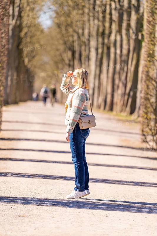 Woman standing in a sunny avenue and looking into the distance. Spring scene with clear tree shadows, Schwetzingen Palace Gardens, Germany