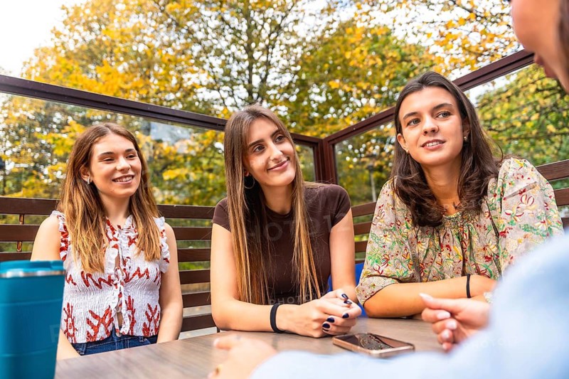 Group of young women friends chatting and laughing at an outdoor cafe terrace on a pleasant autumn day, enjoying coffee, conversation and relaxed togetherness