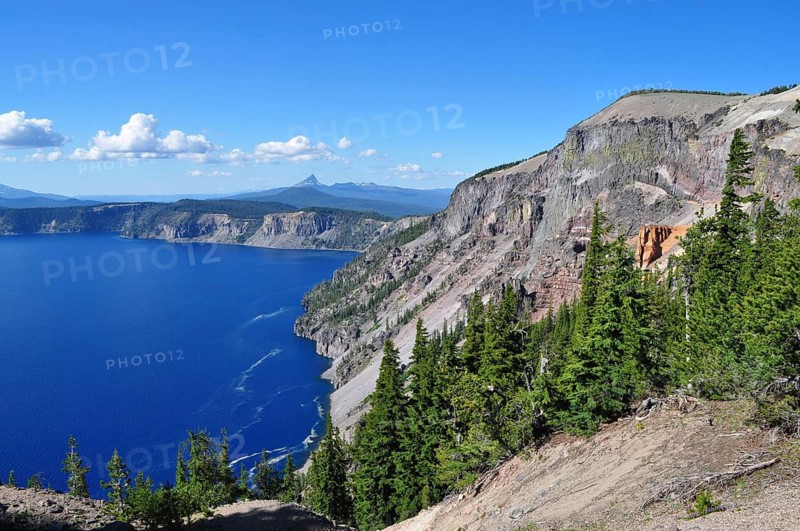 Steep rocks above the deep blue lake with green forest and expansive views of mountains on the horizon, Crater Lake National Park, Oregon, USA