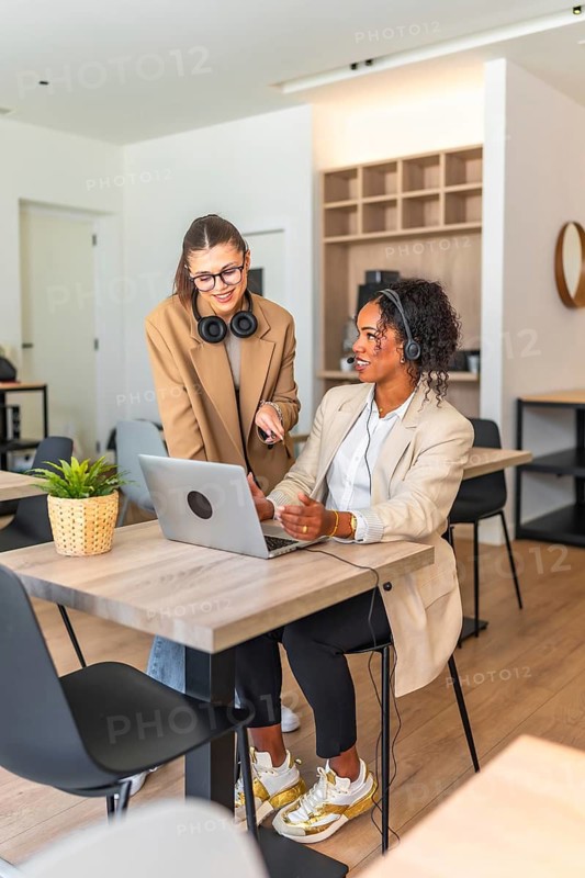 Two businesswomen working together on a laptop, discussing a project in a modern office environment, showcasing teamwork and collaboration