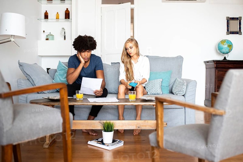 A couple sits on a sofa analyzing financial documents and using a calculator. The setting is a modern, cozy living room with a coffee table and a globe in the background