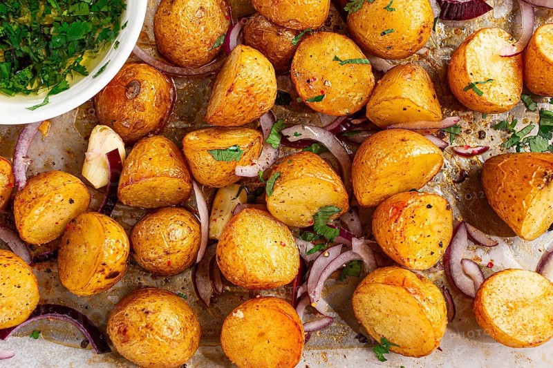Roasted potatoes sit on a baking sheet next to chopped herbs and red onions. They are golden brown and ready to be served. Ingredients add color and flavor to the dish