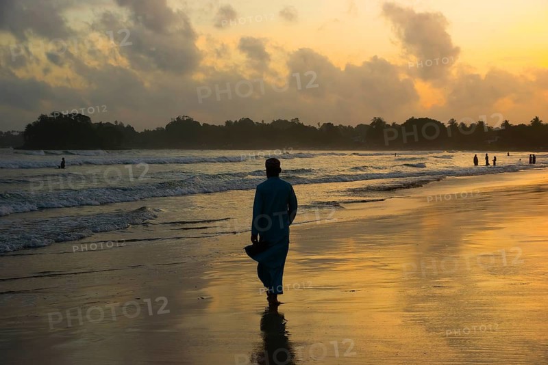 A man strolls along a Sri Lankan beach during sunset, with waves gently rolling in and the sky reflecting warm hues on the wet sand. A tranquil and peaceful scene