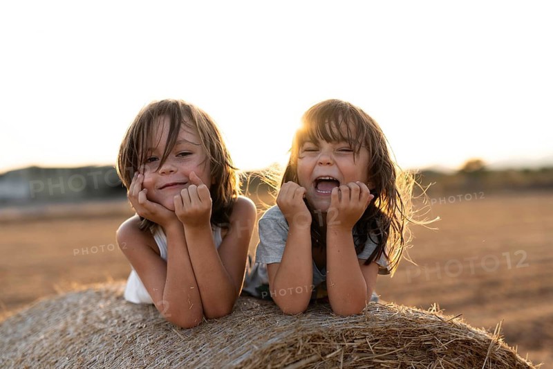 Two happy children lie on a hay bale, bathed in warm sunlight, laughing and enjoying the outdoor setting. Their expressions capture the essence of carefree childhood fun
