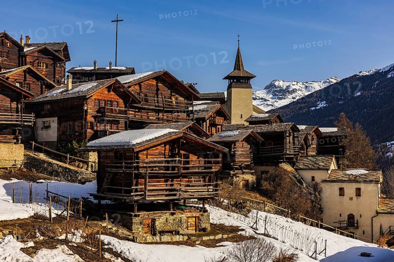 Snow-covered roofs of old farmhouses and barn storage, in the back the wooden cross and the church tower of the Saint Theodule church, historic town center, Grimentz, Val d'Anniviers, Valais Alps, Canton of Valais, Switzerland