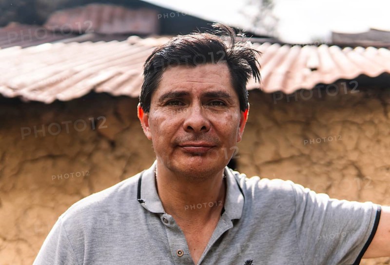 A close-up portrait of a man standing outdoors in a rural area of Colombia, with a rustic clay wall and corrugated metal roof in the background, reflecting a serene expression