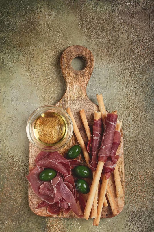 An appetizer of prosciutto, grissini and olives, served on a wooden board, with olive oil