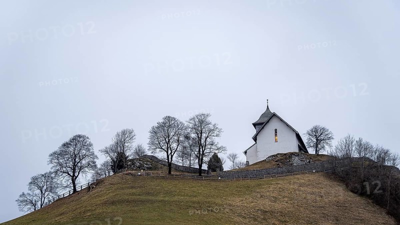 A picturesque white church sits atop a grassy hill, surrounded by bare trees in a hot air balloons festival of castle of Oex. The overcast sky adds a tranquil atmosphere to this countryside scene