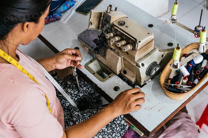 A asian seamstress works at a sewing machine, surrounded by threads and a tape measure. Her focus is on the intricate task, highlighting the craft of garment creation