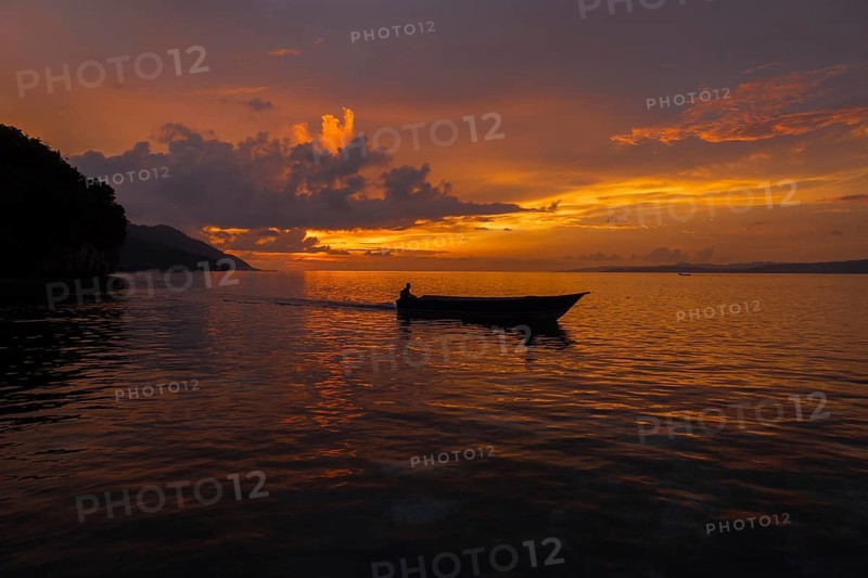 A serene sunset paints the sky in hues of orange and purple over Kri Island, Raja Ampat Archipelago. A small boat and its lone occupant float calmly on the reflective waters, enhancing the tranquil scene