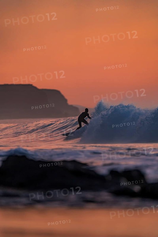 Silhouette of a surfer catching a wave during a vibrant sunset in Fuerteventura, Canary Islands, Spain. The waves crash gracefully, complementing the vivid sky