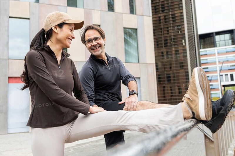 A couple smiles as they engage in an outdoor fitness routine, stretching against a railing. They wear casual sportswear, embodying healthy and active living in an urban setting