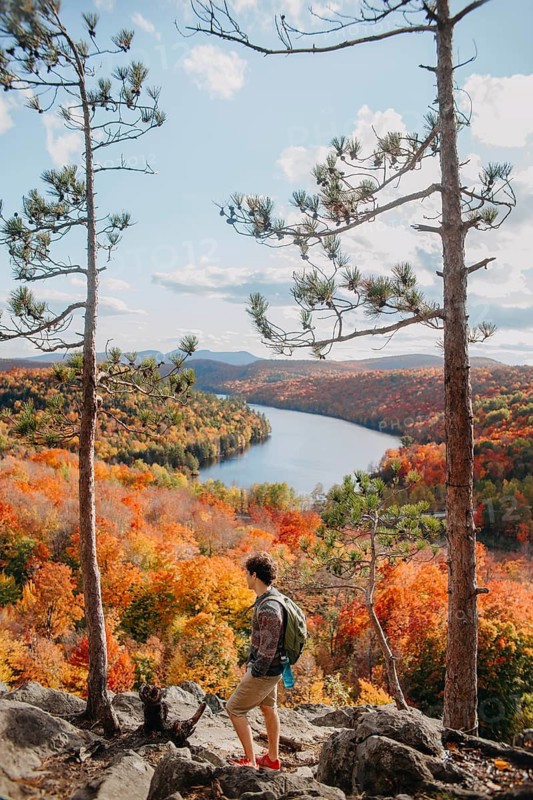 A hiker stands on a rocky outcrop, admiring the stunning autumn foliage and winding river in Quebec, Canada. The vibrant colors create a picturesque and serene atmosphere