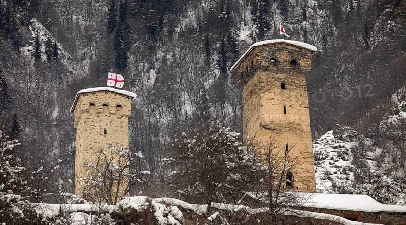 Two ancient stone defense towers stand amid a snowy landscape, with a forested mountain in the background in Georgia One tower displays the Georgian flag, symbolizing historic significance