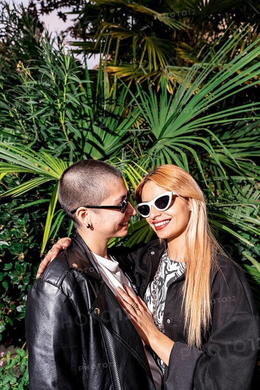 A joyful lesbian couple embraces outdoors, dressed in trendy outfits and sunglasses, standing in front of lush greenery, capturing a moment of love and happiness